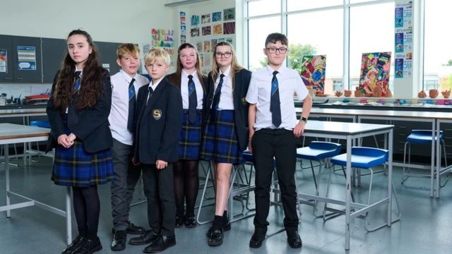The students stand together in the classroom. Three girls and three boys. They are all wearing school uniform and standing in front of a number of desks and stools.