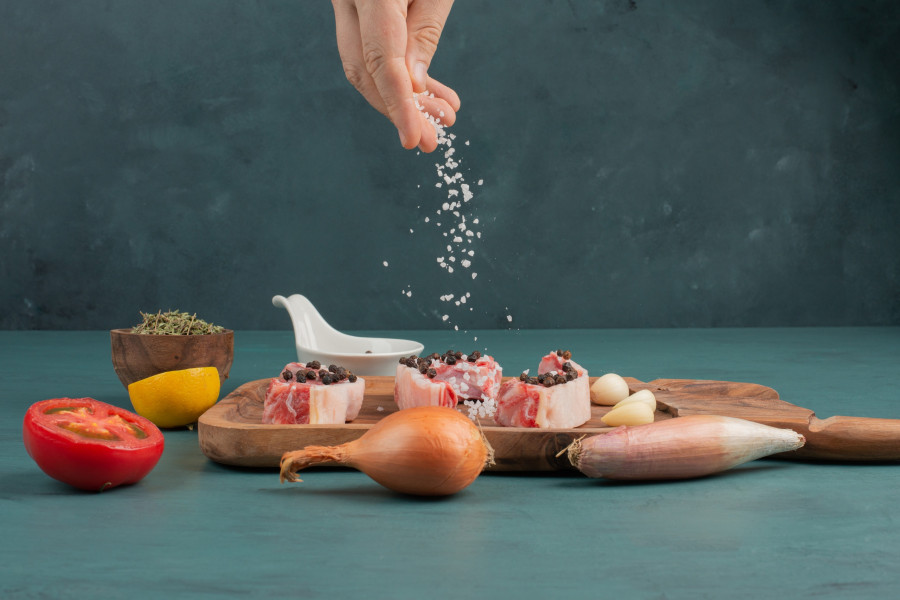 Woman adds salt into uncooked meat pieces on blue background